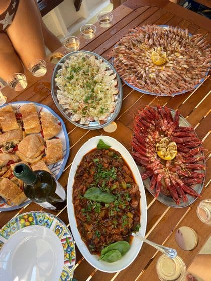 Un pasto all'aperto per un viaggio di gruppo WeRoad con vassoi di gamberi, panini, stufato e vino bianco su un tavolo di legno.