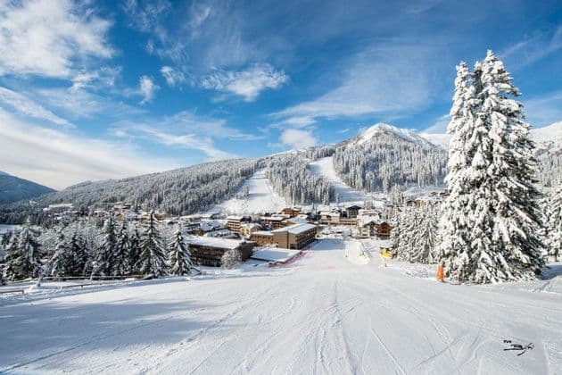 Una pista da sci innevata si affaccia su un villaggio di montagna circondato da foreste di pini sotto un cielo blu con nuvole.