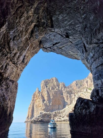 Vista dall'interno di una grotta marina, con una barca su acqua calma e grandi scogliere rocciose sullo sfondo.