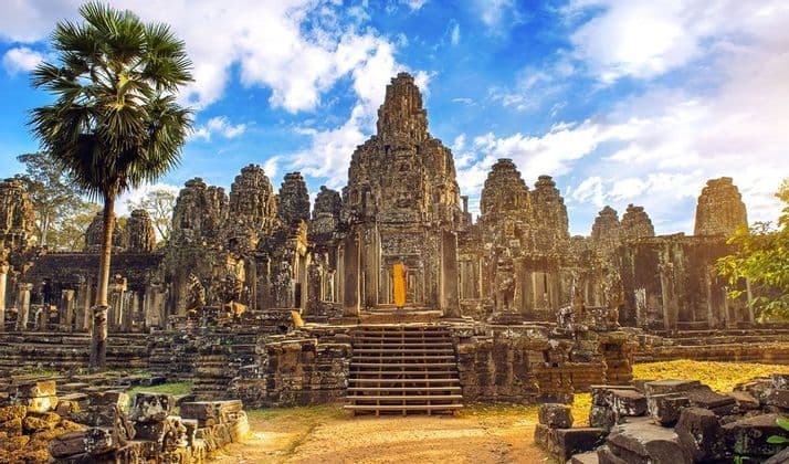 A person in orange robes walks up the stone steps of a large, ancient temple with carved towers, next to a tall palm tree.
