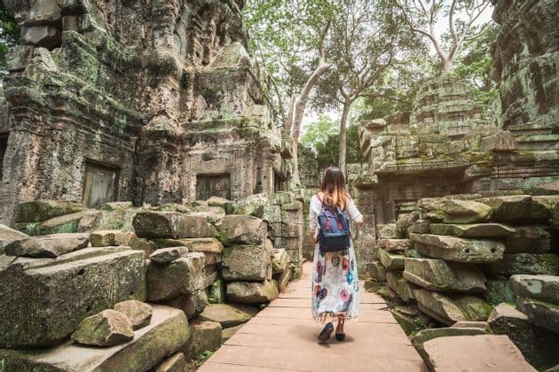 Una mujer con mochila camina por un sendero de madera entre antiguas ruinas de piedra cubiertas de musgo y exuberantes árboles verdes.