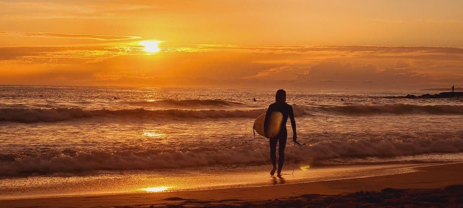 Un surfista in muta trasporta una tavola da surf e cammina lungo la spiaggia verso le onde durante un tramonto arancione splendente.