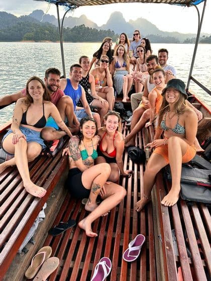 Un grupo de WeRoad en un largo barco de madera, sonriendo a la cámara con un lago y montañas de fondo.