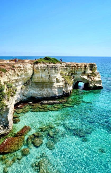 A coastal view of white cliffs and a natural rock arch over clear turquoise water on a sunny day.