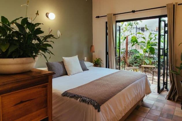 Interior of a bedroom with a double bed, a large potted plant, and open doors leading to a green patio.
