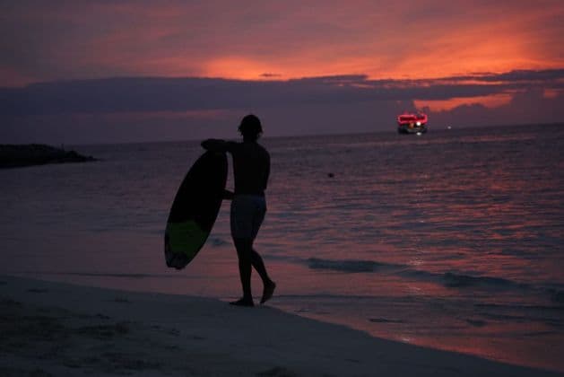 Una persona in silhouette con una tavola da surf cammina lungo la riva al tramonto, con una barca sull'acqua in lontananza.