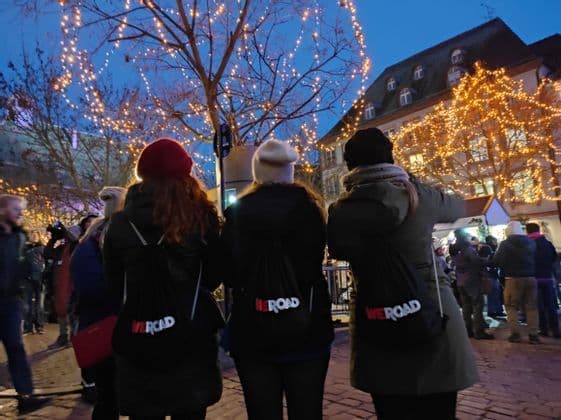 Three people from a WeRoad group trip, seen from behind, stand in a crowded square with trees decorated with festive lights.