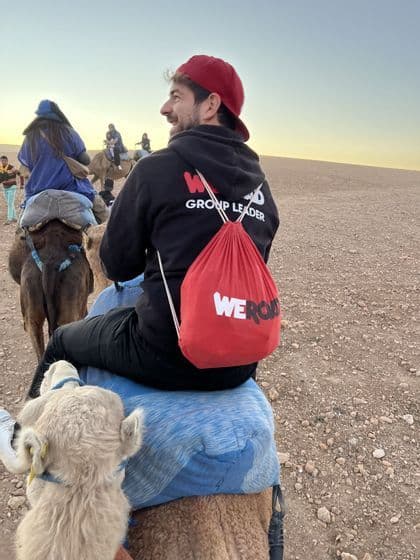 Un accompagnatore WeRoad, visto di spalle, sorride mentre cavalca un cammello nel deserto con altri viaggiatori al tramonto.