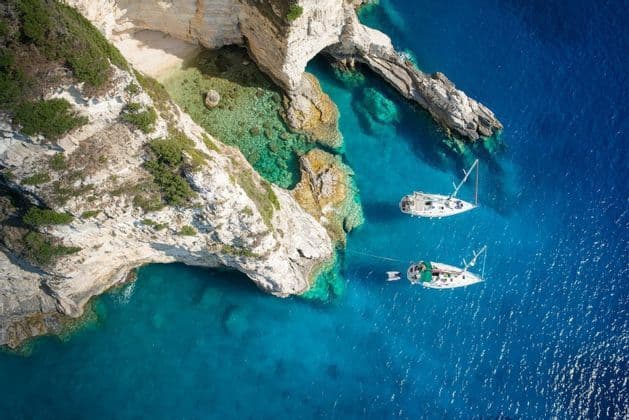 Luftaufnahme von zwei weißen Segelbooten, die im klaren, türkisfarbenen Wasser vor einer felsigen Klippe mit einer kleinen Sandbucht ankern.