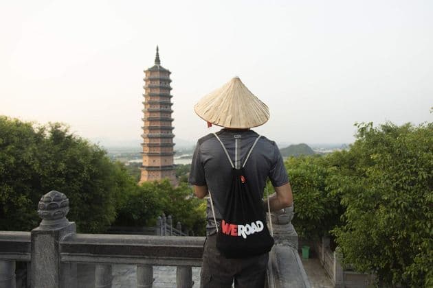 Una persona vista de espaldas, con un sombrero cónico y una mochila WeRoad, contempla una alta pagoda desde un mirador de piedra.
