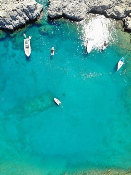 Una vista aerea dall'alto di diverse barche che galleggiano in una cala rocciosa con acqua cristallina turchese.