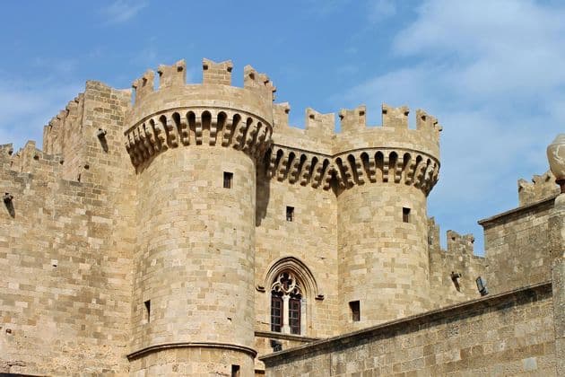 Due grandi torri rotonde di un castello in pietra con merlature, sotto un cielo azzurro con nuvole leggere.