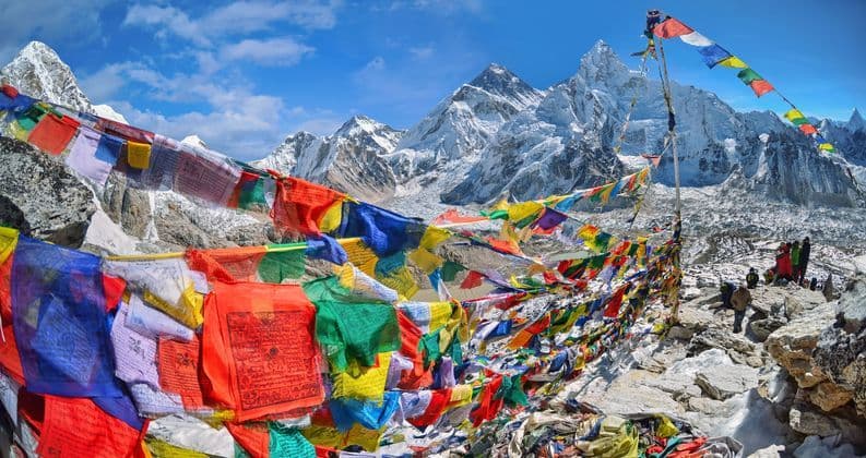 Des drapeaux de prière colorés flottaient sur un point de vue rocheux, avec des montagnes enneigées et un glacier en arrière-plan sous un ciel bleu.