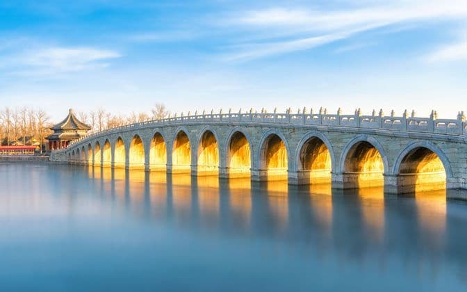 Un lungo ponte di pietra ad arco illuminato dalla luce dorata del sole attraversa acque calme, con il suo riflesso visibile sulla superficie.