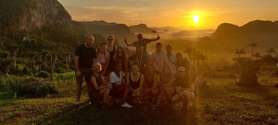 Un groupe WeRoad prend la pose pour une photo sur une colline herbeuse dominant une vallée avec des montagnes et des palmiers au coucher du soleil.