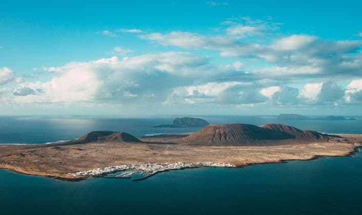 Veduta aerea di un'isola vulcanica con un piccolo villaggio costiero e porto, circondata da un oceano blu intenso sotto un cielo parzialmente nuvoloso.