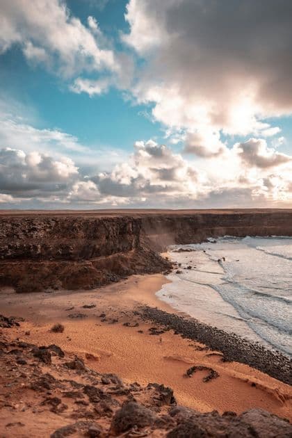 Ein Blick von oben auf eine felsige Küste mit orangem Sand, dunklen Klippen und Meereswellen unter einem teilweise bewölkten Himmel.