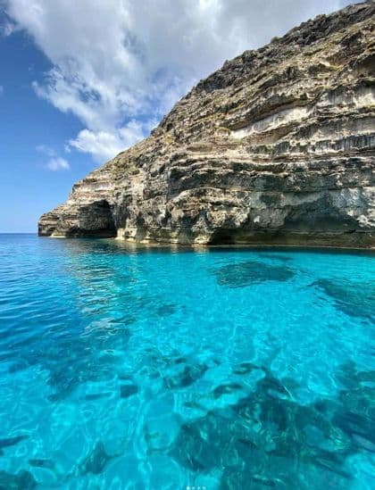 Una vista desde el agua de un acantilado rocoso estratificado que se encuentra con aguas turquesas y cristalinas bajo un cielo parcialmente nublado.