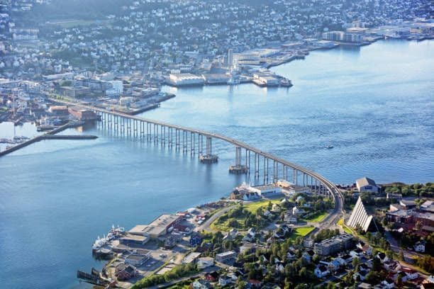Una vista aerea di un lungo ponte che collega una città costiera sull'acqua blu, con un moderno edificio triangolare in primo piano.