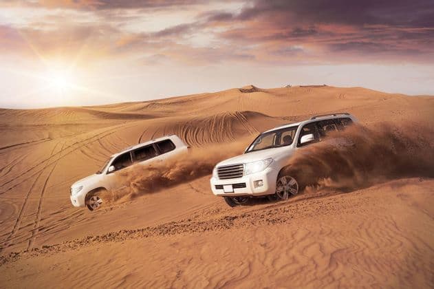 Deux SUV blancs descendent une grande dune de sable dans le désert, soulevant des nuages de sable sous un soleil couchant.