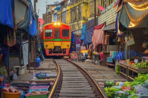Un train rouge et jaune circule sur des rails qui traversent un marché alimentaire extérieur animé, bordé d'étals.