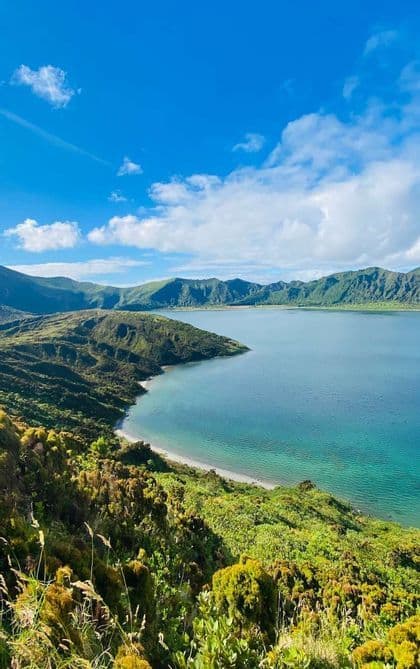 Vista dall'alto di un lago craterico turchese, circondato da lussureggianti montagne verdi, sotto un cielo azzurro e nuvoloso.