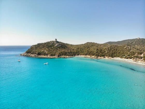 Vista aerea di una baia costiera con acqua turchese vivace, una spiaggia di sabbia e una torre di avvistamento in pietra su una collina verde.
