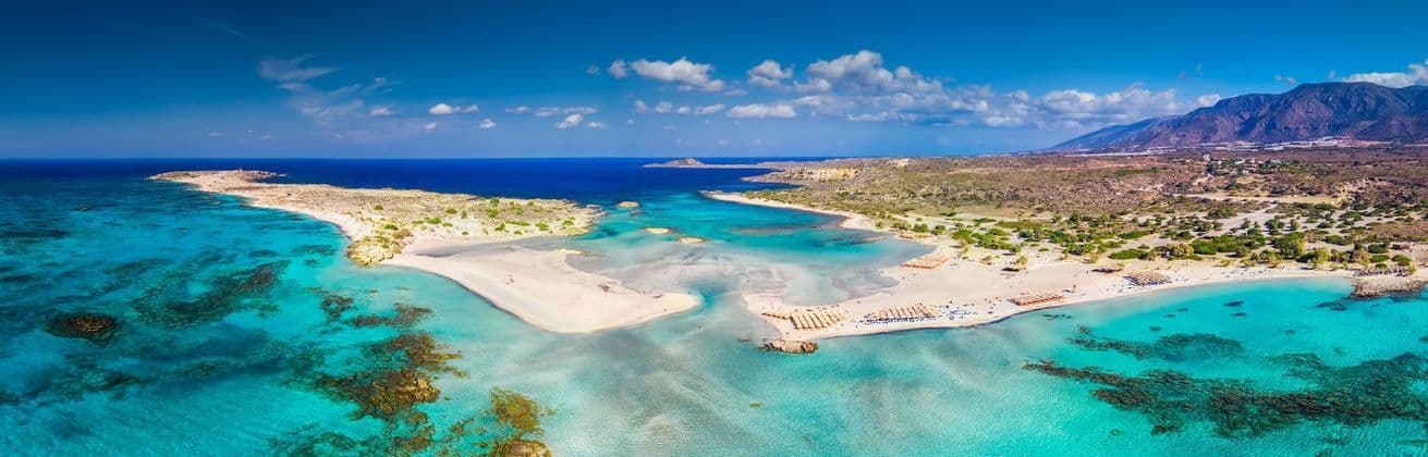 Aerial view of a sandy peninsula stretching into shallow turquoise water, with mountains on the distant coastline under a blue sky.