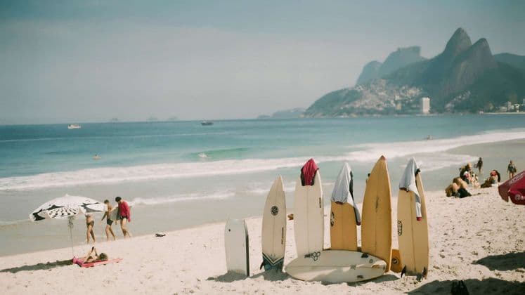 Diverse tavole da surf sono in piedi sulla sabbia di una spiaggia assolata, con persone che si rilassano vicino all'oceano e montagne sullo sfondo.
