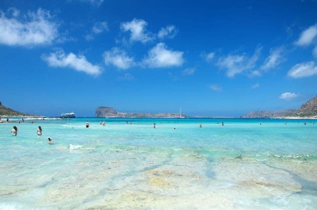 People swimming and relaxing in shallow, clear turquoise water at a beach, with rocky islands under a blue sky with white clouds.