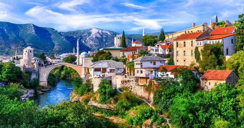 A coastal town with red-tiled roofs and a stone bell tower on the edge of a turquoise bay, with mountains in the distance.