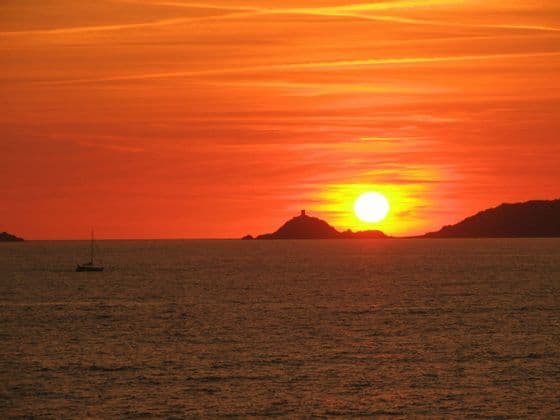 Il sole tramonta sull'oceano, stagliando un'isola con una torre e una barca a vela sull'acqua, tutto sotto un cielo arancione.