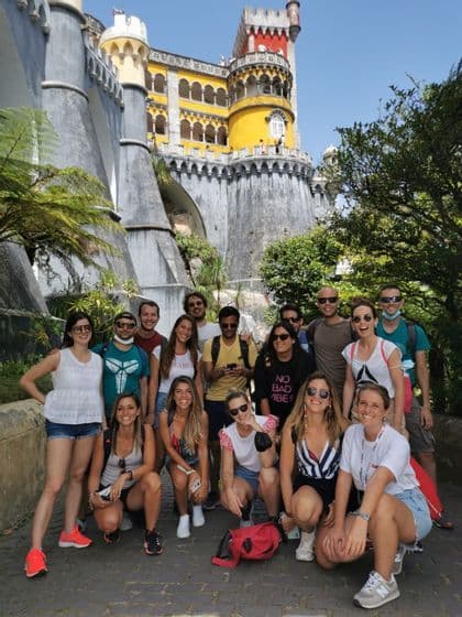 Un viaje en grupo de WeRoad sonríe para una foto grupal frente a un colorido castillo amarillo y gris bajo un cielo azul claro.