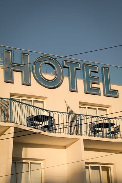 Una vista dal basso della facciata di un hotel con una grande insegna blu retrò sopra balconi con tavoli e sedie sotto un cielo sereno.