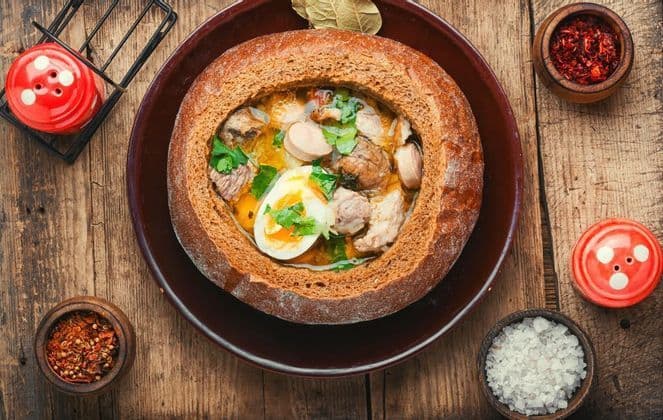 Eine Draufsicht auf eine Suppe mit Fleisch, Wurst und einem hartgekochten Ei, serviert in einem ausgehöhlten Brotleib auf einem Holztisch.