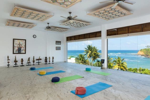 Ein helles, leeres Yogastudio mit Matten und Kissen auf einem Marmorboden, mit großen Fenstern, die einen tropischen Ozean und Palmen zeigen.