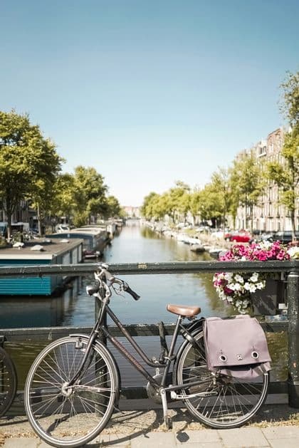 Una bicicleta negra con una bolsa trasera está estacionada en un puente con vistas a un canal bordeado de casas flotantes, árboles y edificios.
