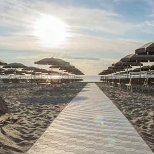 Una passerella piastrellata su una spiaggia sabbiosa conduce tra file di sedie e ombrelloni vuoti verso il mare al tramonto.