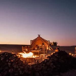 Un falò arde in una buca rivestita di pietre, davanti a una grande tenda illuminata in un campeggio nel deserto, al tramonto.