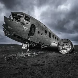 Das Wrack eines Flugzeugrumpfes liegt auf einem Feld aus schwarzem Sand unter einem stark bewölkten Himmel.