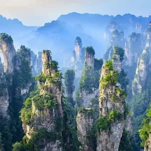 Alte formazioni rocciose a pilastro coperte di vegetazione verde si ergono da una valle nebbiosa con montagne sullo sfondo.