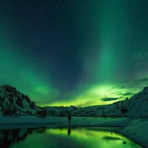 Eine Person beobachtet die Nordlichter über schneebedeckten Bergen, deren grünes Leuchten sich im ruhigen Wasser eines Sees spiegelt.