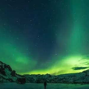 Una persona de pie en una orilla nevada, observando la aurora boreal verde reflejarse en el agua bajo un cielo estrellado.