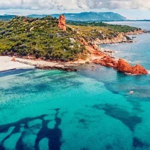 Vista aerea di una costa con acqua turchese, una spiaggia di sabbia bianca, rocce rosse e colline verdi sotto un cielo nuvoloso.