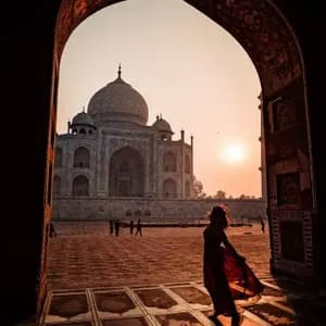 Una donna in un fluente abito rosso è in silhouette in un arco scuro, incorniciando una vista del Taj Mahal all'alba.