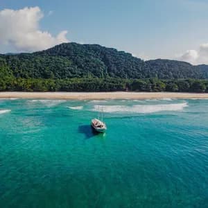 Vue aérienne d'un voilier blanc ancré dans des eaux turquoise, avec une plage de sable et une montagne verdoyante en arrière-plan.