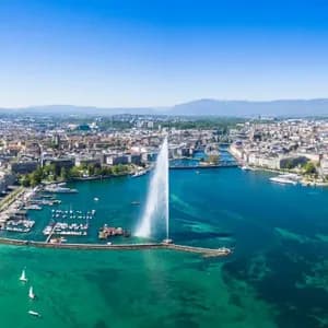 Vista aerea di una grande fontana che zampilla acqua da una baia turchese, con un denso panorama urbano e montagne distanti.