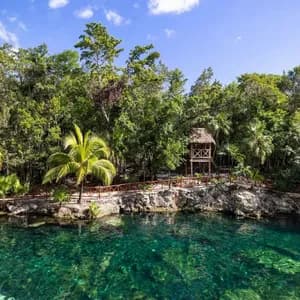 Un cenote limpido color turchese delimitato da una lussureggiante giungla tropicale con una torre di avvistamento in legno sotto un cielo parzialmente nuvoloso.