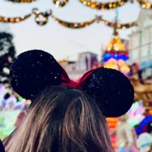 La nuca di una persona che indossa orecchie da topo nere con paillettes, con una parata sfocata e festosa sullo sfondo.