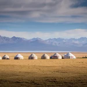 Une rangée de yourtes dans une plaine herbeuse, face à un lac et une chaîne de montagnes lointaine.